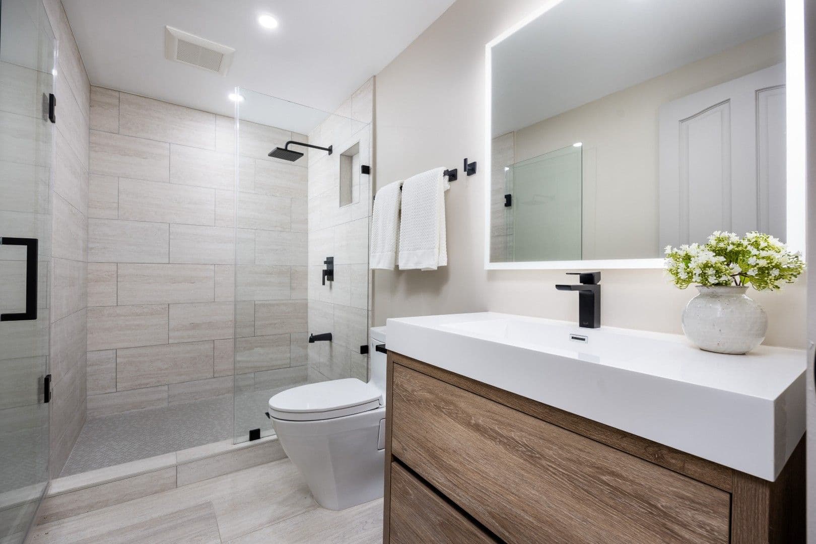 Modern bathroom with walk-in shower, white rectangular sink, backlit mirror, wooden vanity, toilet, and decorative plant in vase