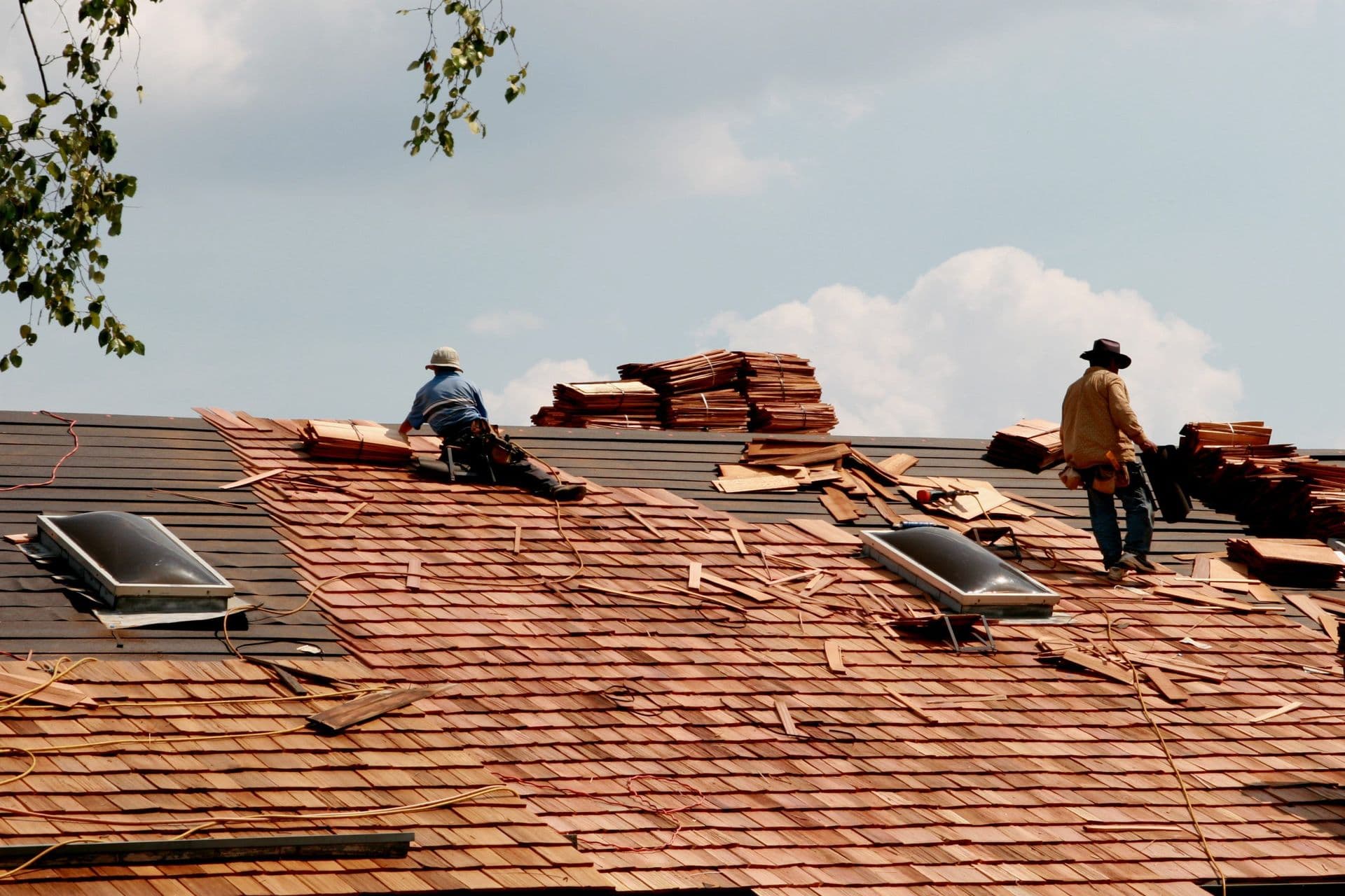 Two workers install wooden shingles on a sloped roof under a sunny sky. They wear safety gear, surrounded by stacks of shingles and tools.