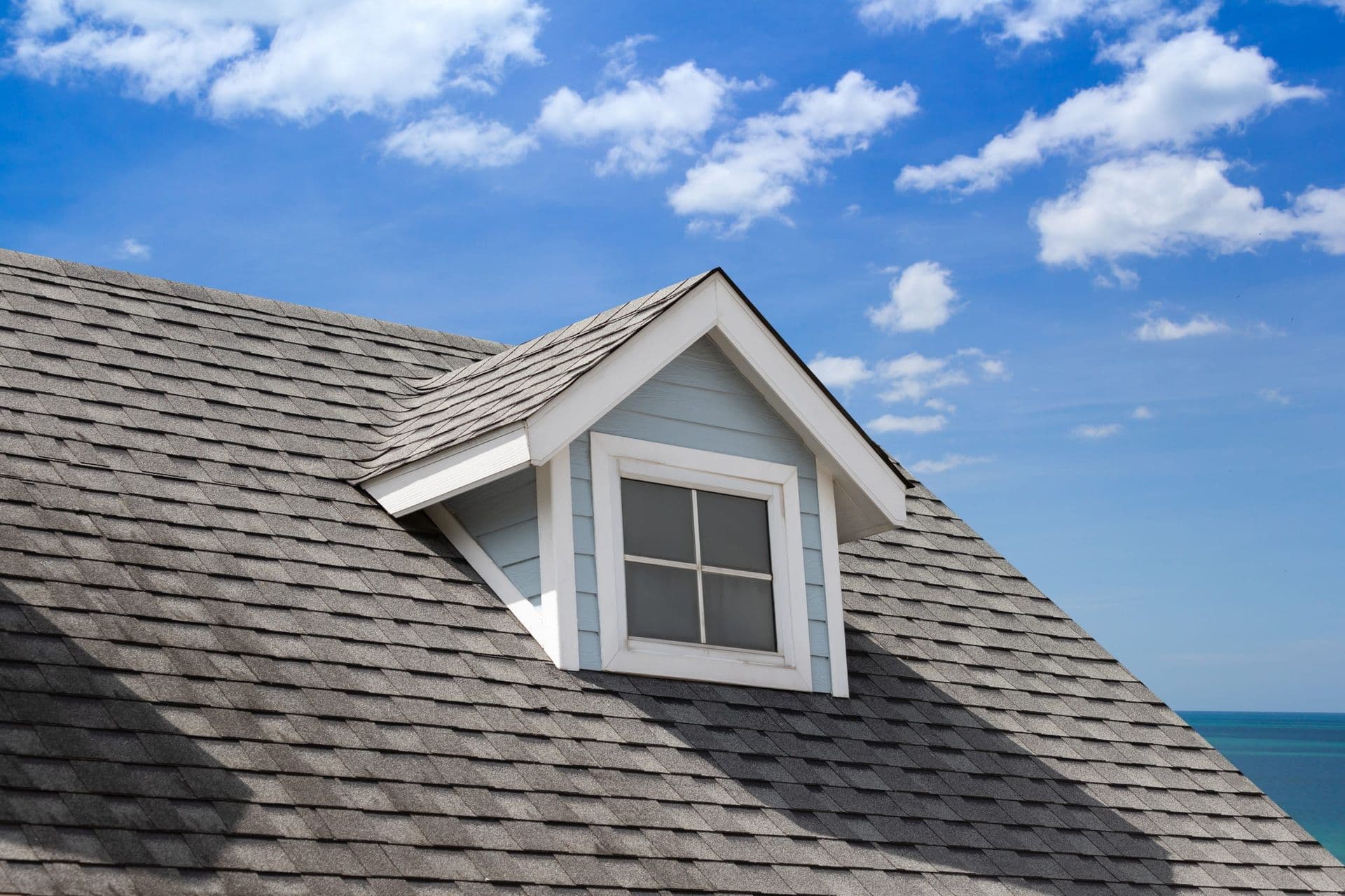 A gray shingled roof with a small dormer window under a bright blue sky with scattered clouds. The ocean is visible in the background, suggesting a coastal setting.